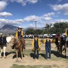 Students dressed in blue and gold western wear. Two students are atop horses and two students are cheering them on by holding pom poms.